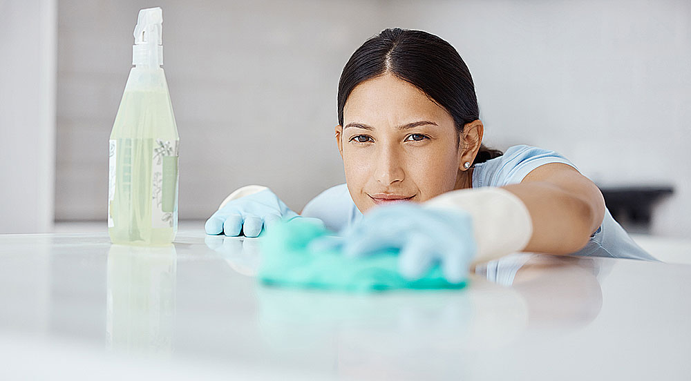 Cleaner in blue gloves wiping a glossy countertop until it shines, spray bottle beside her